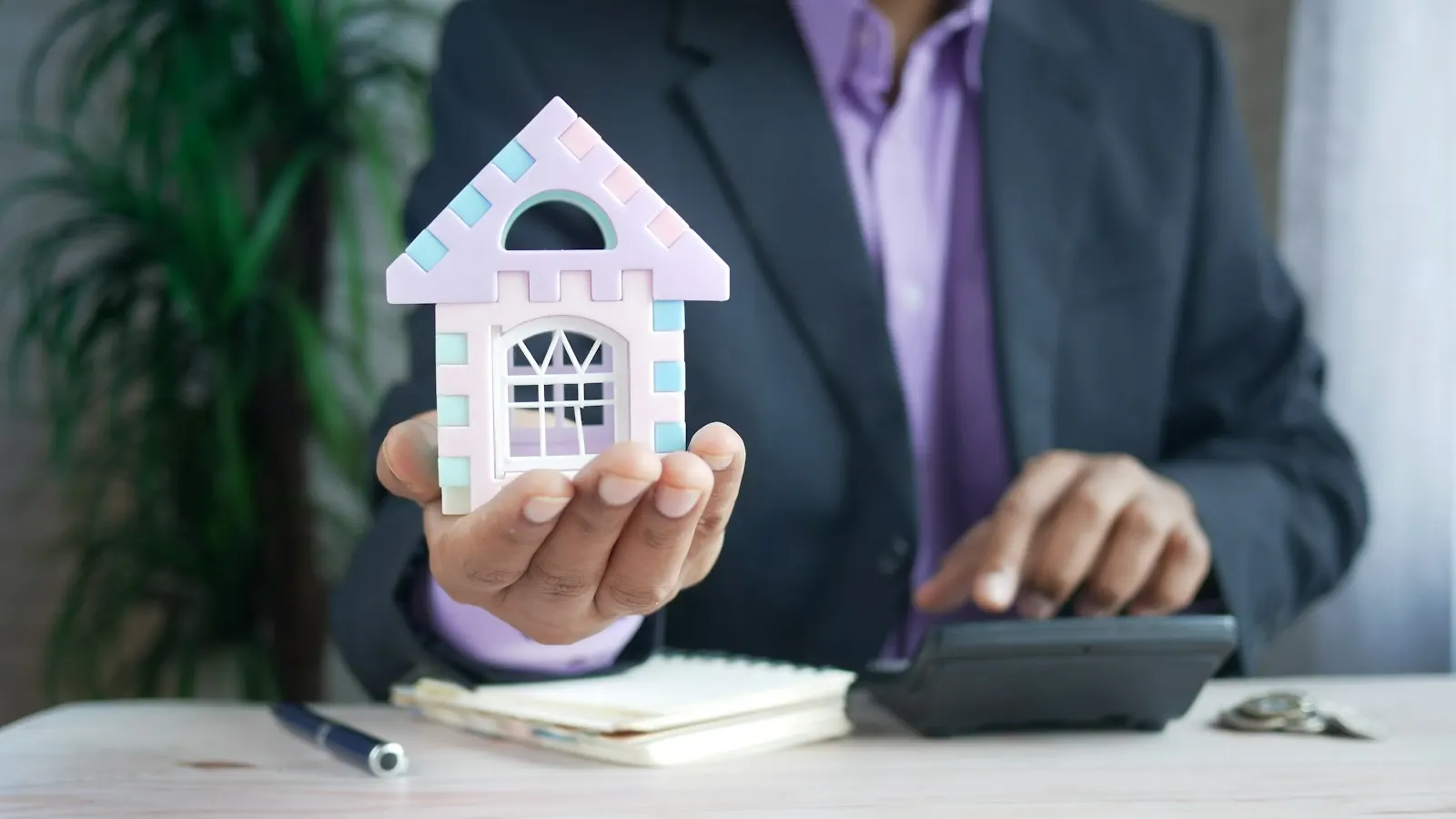 A man holding a small model house at a desk with a calculator